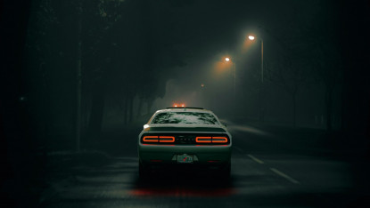 A green car drives on a foggy, dimly-lit road at night. Red brake lights glow, while streetlights cast a soft light. The scene feels mysterious and atmospheric.