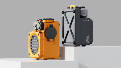 Two portable speakers on white blocks: one bright orange with black accents, the other sleek black with a gray knob. Modern, industrial design.