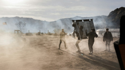 A group of men maneuvers a large netting amidst a dusty, desert landscape. A tractor and American flag are visible in the background, under a hazy sky.