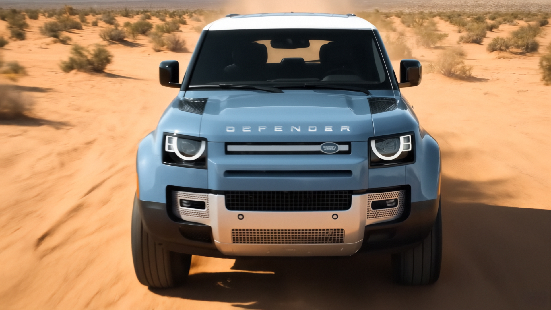 A blue SUV with "Defender" on the hood drives on a sandy desert road with sparse bushes. The sky is clear, conveying an adventurous tone.