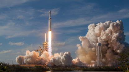 A rocket launches into a blue sky, leaving behind a trail of bright flame and thick white smoke, conveying energy and excitement.