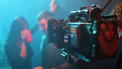 Film set with a focus on a large camera in dim lighting. Out-of-focus figures, one with a red cup, suggest a casual, behind-the-scenes atmosphere.