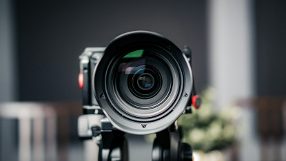 Close-up of a camera lens with a blurred background. The lens is centered, capturing the scene with precision. The tone is professional and focused.
