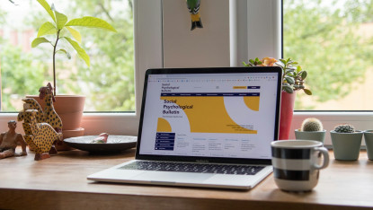 A laptop displaying a website titled "Social Psychological Bulletin" is on a wooden desk. Plants, a striped mug, and decorative figurines surround it, creating a cozy, vibrant workspace.